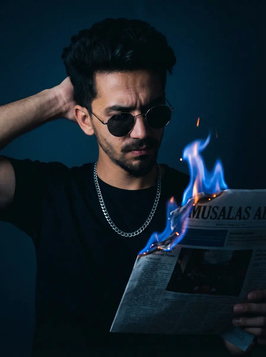 Close-up cinematic portrait of a young man in a black t-shirt thick silver chain and round sunglasses seriously reading a Musalas AI newspaper with the top right corner burning in dramatic bright blue flames and orange embers under moody dark blue low key lighting with one hand placed behind his head